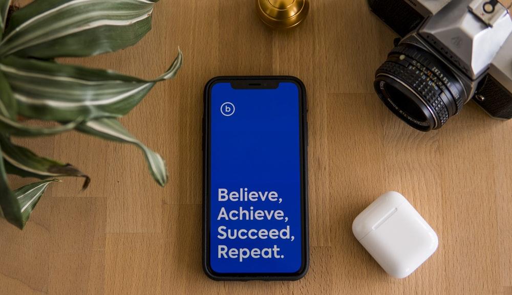 Smartphone lying on table; the screen has the words "believe, achieve, succeed, repeat".