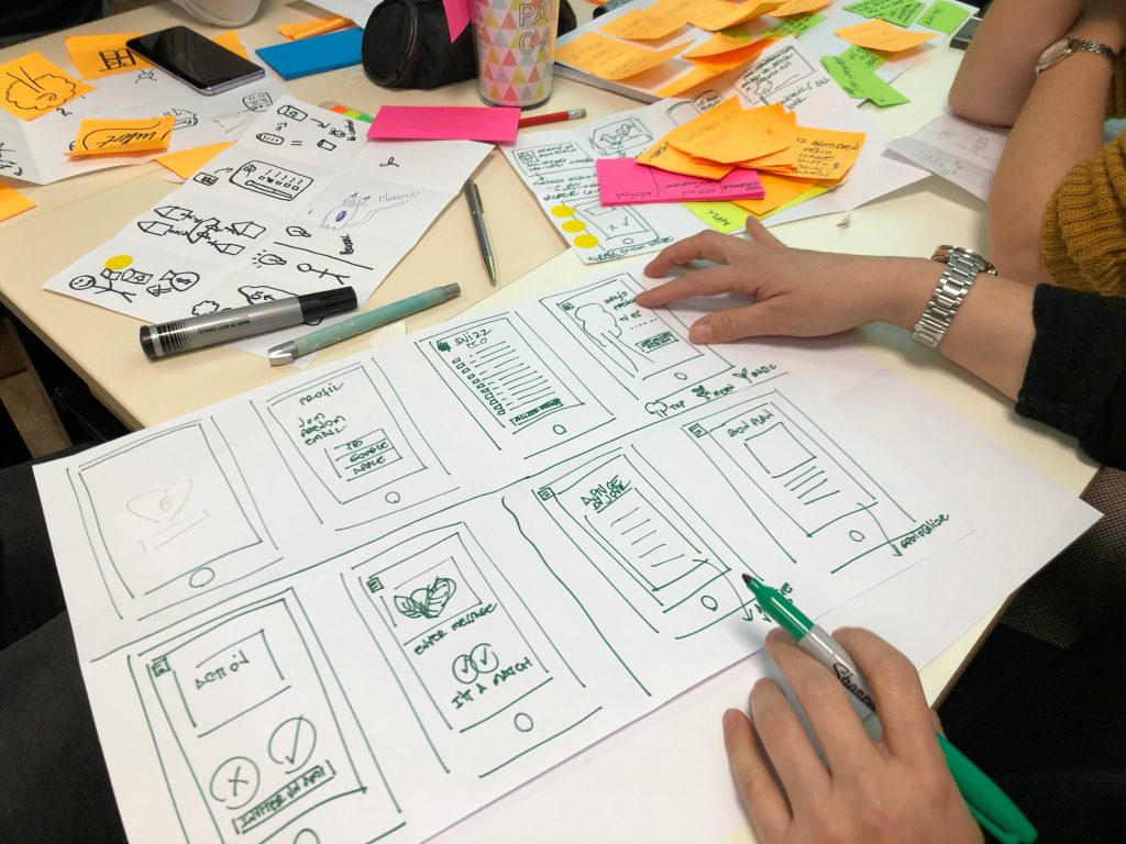 Several people off screen are working on the UX design of an app. We can see their hands and the papers on which they have drawn wireframes and visual aids to better understand their users.